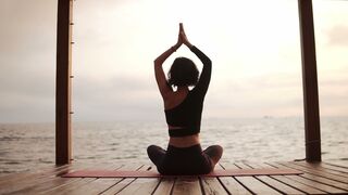 Woman practicing yoga and stretching spine and hands sitting on mat on sea pier holding hands above